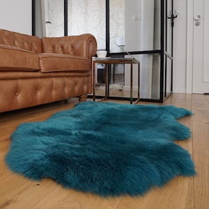 May include: A plush, deep teal-colored sheepskin rug with a thick, shaggy texture and an irregular, organic shape. The rug features long, soft fibers that create a dense pile, providing a rich, saturated color that contrasts with the wooden floor.