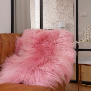 May include: A plush, long-pile faux fur throw rug in a vibrant shade of pink. The material features a dense, shaggy texture with soft, flowing fibers that drape over the surface of a brown leather sofa, creating a thick and layered appearance.