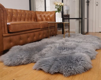 May include: A large, rectangular, grey sheepskin rug with a thick, long-pile texture. The rug features a natural, irregular edge shape and a dense, soft appearance, covering a portion of the wooden floor in front of a brown leather sofa.