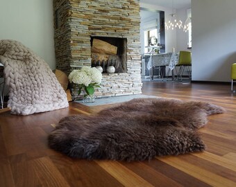 May include: A dark brown sheepskin rug with a thick, textured fleece surface. The rug has an irregular, natural shape and is positioned on a wooden floor. The fur appears dense and soft, covering the floor area in front of a stone fireplace.
