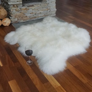May include: A plush, off-white sheepskin rug with a thick, long-pile texture and irregular, natural edges. A clear glass stemware containing a dark red liquid is positioned on the floor next to the rug, providing a sense of scale for the item.