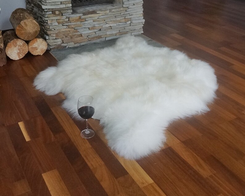 May include: A plush, off-white sheepskin rug with a thick, long-pile texture and irregular, natural edges. A clear glass stemware containing a dark red liquid is positioned on the floor next to the rug, providing a sense of scale for the item.