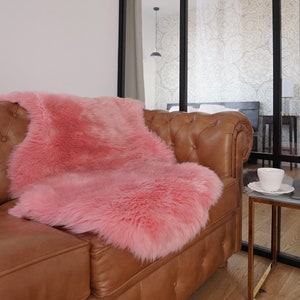 May include: A soft, fluffy, pink faux fur rug with a long-pile texture draped over a brown leather tufted sofa. The rug has an irregular, organic shape and a vibrant, solid pink color that contrasts with the smooth, tan-colored leather upholstery.