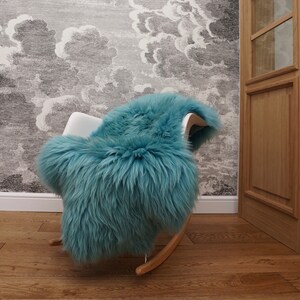 May include: A plush, teal-colored sheepskin rug draped over a white rocking chair with wooden runners. The rug features long, thick, and soft fur fibers that cover the seat and backrest of the chair, creating a dense and textured appearance.