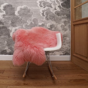 May include: A pink faux fur rug draped over a white rocking chair. The chair is in front of a wall with a gray and white cloud pattern.