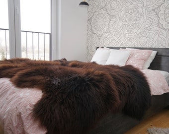 Op de afbeelding: Een bruine schapenvacht deken gedrapeerd over een bed met witte en roze beddengoed. Het bed staat tegen een muur met een wit en grijs patroon behang.
