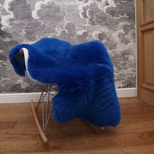 May include: A vibrant royal blue sheepskin rug draped over a white rocking chair with a chrome wire base and light wood rockers. The thick, plush fur texture covers the seat and backrest of the chair, creating a soft and dense appearance.