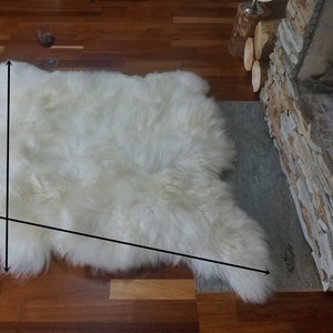 May include: A white sheepskin rug with long, dense, and soft fur. The rug has an irregular, natural shape with visible black arrows indicating the length and width dimensions across the surface of the white, fluffy, and textured material.