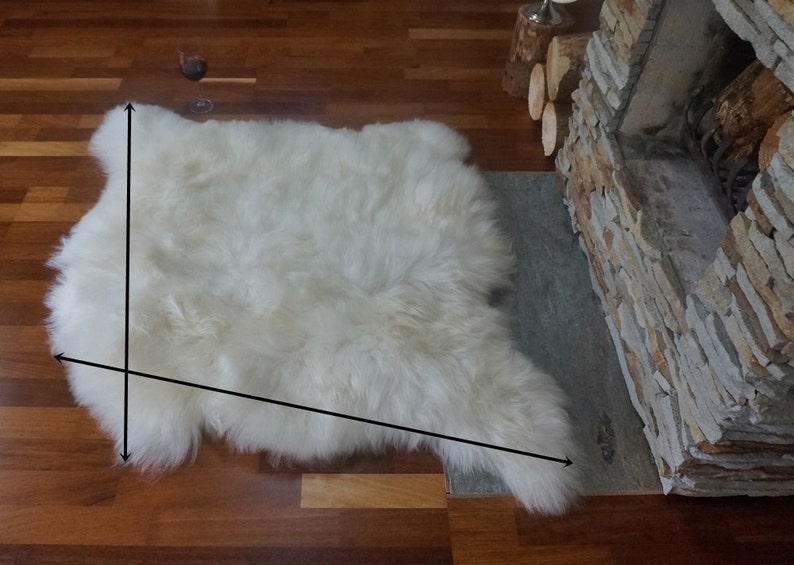 May include: A white sheepskin rug with long, dense, and soft fur. The rug has an irregular, natural shape with visible black arrows indicating the length and width dimensions across the surface of the white, fluffy, and textured material.