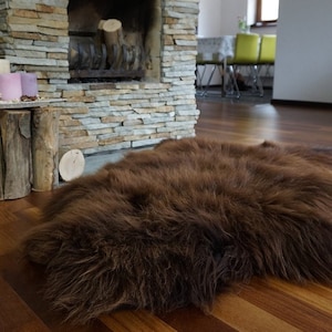 May include: A brown sheepskin rug with long, soft fur. The rug is laid out on a hardwood floor in a living room setting.