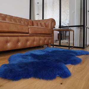 May include: A vibrant royal blue sheepskin rug with a thick, plush texture and an irregular, natural organic shape. The rug features dense, soft fur fibers that create a deep, saturated color profile, laid flat on a light-toned wooden floor.