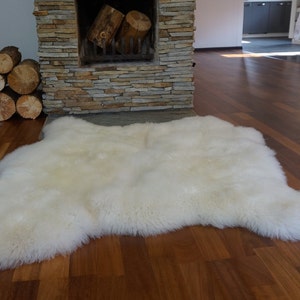 May include: A large, off-white sheepskin rug with a thick, dense, and fluffy texture. The rug features an irregular, natural hide shape with soft, long fibers that create a plush appearance. The surface is uniform in color, showcasing a creamy tone.