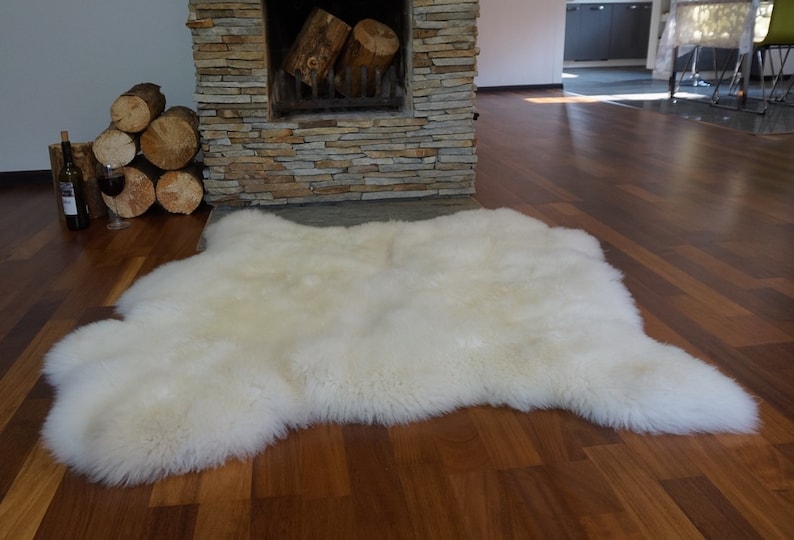 May include: A large, off-white sheepskin rug with a thick, dense, and fluffy texture. The rug features an irregular, natural hide shape with soft, long fibers that create a plush appearance. The surface is uniform in color, showcasing a creamy tone.