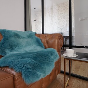 May include: A plush, teal-colored faux fur throw blanket draped over a brown leather tufted sofa. The throw has a thick, shaggy texture and covers a significant portion of the seat and backrest of the sofa, providing a soft contrast to the leather.