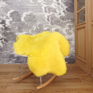 May include: A bright yellow faux fur sheepskin rug draped over a rocking chair. The rug features a thick, plush texture and a vibrant, solid yellow color. The rocking chair has a white seat shell, thin metal supports, and light wood rockers.