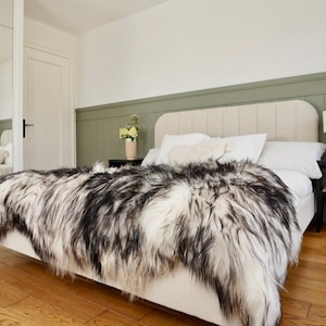 Pode incluir: Uma cama com cabeceira creme e uma manta de pele sintética preta e branca. A cama é feita com lençóis e almofadas brancas. Uma pequena lâmpada de madeira está em uma mesa de cabeceira preta. O quarto tem piso de madeira e uma parede verde.