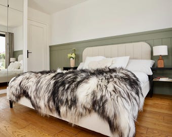 May include: A bed with a cream-colored headboard and a black and white faux fur throw. The bed is made up with white sheets and pillows. A small wooden lamp sits on a black nightstand. The room has hardwood floors and a green accent wall.