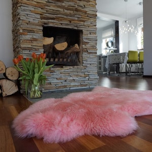 May include: A soft, plush sheepskin rug in a vibrant pink hue with a natural, irregular shape and thick, dense fur texture. The rug is laid out flat on a dark wooden floor, showcasing its long, fluffy pile and consistent, solid pink color throughout.