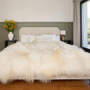 May include: A bed with a cream-colored headboard and a large, fluffy, off-white faux fur blanket. The room has a neutral color palette, with a vase of yellow flowers on a side table. A window with a view is visible.