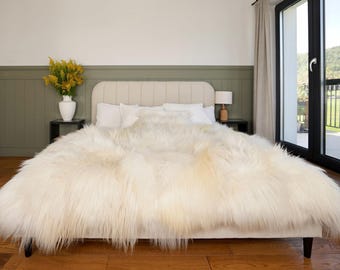 May include: A large, off-white, long-haired sheepskin throw blanket draped over a bed. The textile features a thick, shaggy texture with natural variations in color, covering the entire surface of the mattress and extending over the edges.