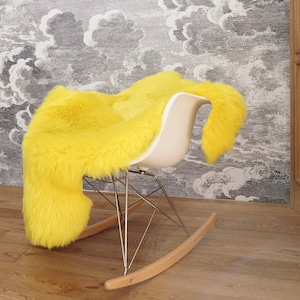 May include: A bright yellow sheepskin rug draped over a white molded plastic rocking chair. The chair features a curved seat shell, a chrome metal wire base, and light-colored wooden rocker runners that sit on the floor.