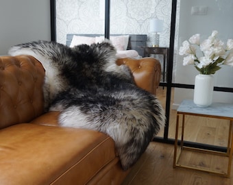 May include: A long-haired sheepskin rug with a gradient of black, grey, and white fur draped over a tan leather tufted sofa. The rug features a thick, shaggy texture with dark charcoal tips transitioning into a lighter, cream-colored base.