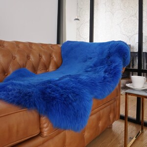 May include: A vibrant royal blue sheepskin rug with a thick, plush texture. The rug is draped over a tufted tan leather sofa, showcasing its long, soft fur fibers and deep, saturated color that covers the seat and backrest of the furniture piece.