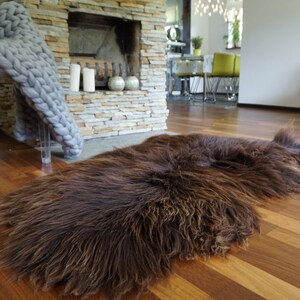 May include: A brown sheepskin rug with long, shaggy fur. The rug is laid out on a hardwood floor in a living room setting.