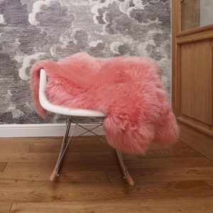 May include: A plush, long-haired pink sheepskin rug draped over a white molded plastic rocking chair with a chrome wire base and light wood runners. The rug features a dense, curly texture and a vibrant, solid pink color throughout its surface.