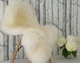 May include: A plush, off-white sheepskin rug with long, dense fur fibers. The rug is draped over a chair, showcasing its soft, thick texture and natural, irregular shape. The material appears fluffy and voluminous, providing a thick layer of padding.