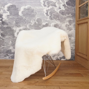 May include: A white sheepskin rug draped over a rocking chair. The chair features a molded seat, a chrome metal wire base, and light-colored wooden rockers. The thick, soft, and fluffy texture of the white fur covers the entire seat area.