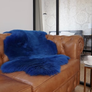 May include: A vibrant royal blue faux fur throw blanket with a thick, shaggy texture draped over a brown leather tufted sofa. The blanket features an irregular, organic shape with long, dense fibers that create a soft and plush appearance.