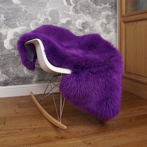 May include: A vibrant purple sheepskin rug draped over a white molded plastic rocking chair. The chair features a chrome metal wire base and light wood rockers. The long-pile, plush purple fur covers the seat and backrest of the chair.
