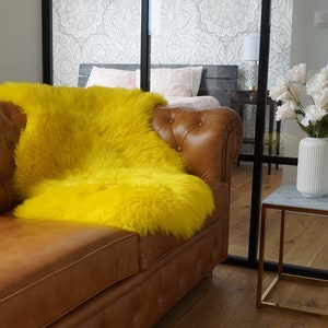 May include: A bright yellow faux fur throw blanket draped over a brown leather tufted sofa. Next to the sofa is a white ribbed ceramic vase filled with white flowers, sitting on a square side table with a marble top and a gold metal frame.