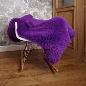 May include: A vibrant purple sheepskin rug draped over a white molded plastic rocking chair with a chrome wire base and light wood runners. The thick, long-pile fur covers the seat and backrest, creating a soft, textured appearance on the chair.