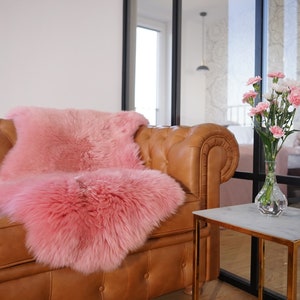 May include: A fluffy, bright pink sheepskin rug draped over a tan leather tufted sofa. The rug features a thick, long-pile texture and a vibrant pink color that contrasts with the smooth, brown leather surface of the button-tufted furniture piece.
