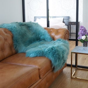 May include: A tufted tan leather sofa featuring a long-haired teal faux fur throw blanket draped over the back and seat cushions, positioned next to a small square side table with a white marble top and a gold metal frame holding a vase of flowers.