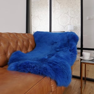 May include: A vibrant royal blue sheepskin rug with a thick, plush texture draped over a tan tufted leather sofa. The rug features long, dense fibers and a deep, saturated blue color that contrasts with the warm brown tones of the leather furniture.