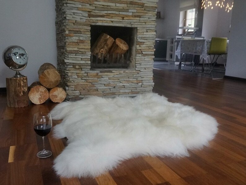 May include: A plush, white sheepskin rug with a thick, long-pile texture. The rug features an irregular, natural shape with soft, dense fibers that create a fluffy appearance. It is positioned on a dark wooden floor near a stone fireplace.