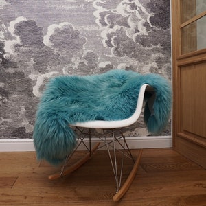 May include: A white rocking chair with a chrome metal base and wooden rockers, draped with a long-haired teal faux fur throw. The chair features a molded seat and a wire frame support structure, providing a contrast to the soft, textured teal fur.