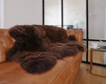 May include: A dark brown, long-haired sheepskin rug draped over a tan leather tufted sofa. The sheepskin has a thick, dense, and shaggy texture with natural variations in the deep brown fur, providing a soft and plush appearance on the seat.
