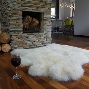 May include: A large, off-white, plush sheepskin rug with a natural, irregular shape. The rug features a thick, soft, and dense pile texture, providing a fluffy appearance. It is laid flat on a dark brown wooden floor, showcasing its organic form.
