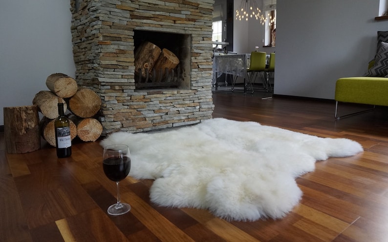 May include: A large, off-white, plush sheepskin rug with a natural, irregular shape. The rug features a thick, soft, and dense pile texture, providing a fluffy appearance. It is laid flat on a dark brown wooden floor, showcasing its organic form.