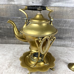 May include: Antique brass tea kettle with a black handle and ornate detailing. The kettle sits on a matching stand with decorative legs and a scalloped edge. A small lid is also visible.