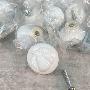 May include: A collection of round white ceramic cabinet knobs, each featuring a raised embossed pear design on the front surface. Several knobs are individually wrapped in clear plastic bags, and one metal mounting screw is placed separately.