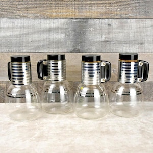 May include: Set of four glass pitchers with black and silver striped bands and black handles. The pitchers are clear glass and have a round shape.