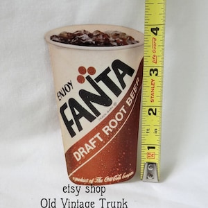 May include: Vintage Fanta Draft Root Beer cardboard cutout, featuring a red and white cup with ice cubes and the Fanta logo. The text "Enjoy" is printed on the cup, along with the words "Draft Root Beer" and "a product of The Coca-Cola Company".