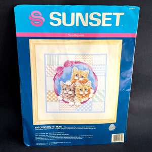May include: A Sunset needlepoint kit titled "Patchwork Kittens." The kit features a cross-stitch design of three kittens in a blue circle, surrounded by a patchwork background. The packaging is blue with the Sunset logo and the text "Needlepoint."