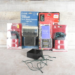 May include: Four packages of battery-operated Christmas lights. The packages are red and white with green and red lights. The lights are on a white wooden surface. The packages have the text "CLEAR best value 10-ct. Battery Operated Light Set" and "10 Life Battery Operated Light Set".
