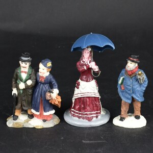 May include: Miniature figurines of people in Victorian-era clothing. One couple is arm-in-arm, another figure holds a blue umbrella, and a third figure wears a blue coat and carries a book. The figurines are on a white base.